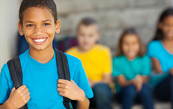 Boy in blue shirt smiling wearing a black strapped schoolbag.